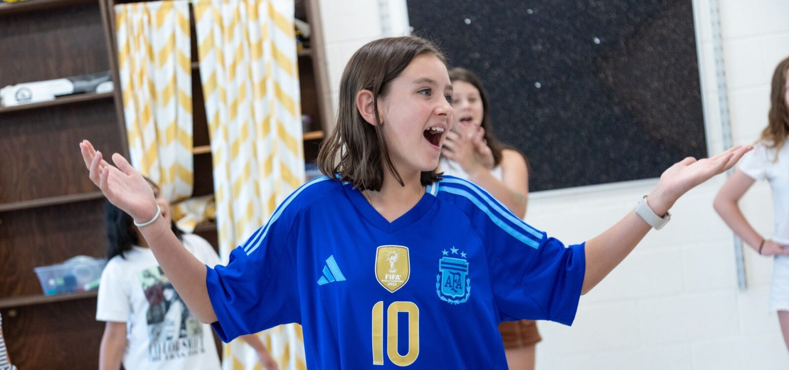 A girl wearing a blue Argentina soccer jersey with the number 10 stands indoors with her arms raised and smiling. Others are blurred in the background.