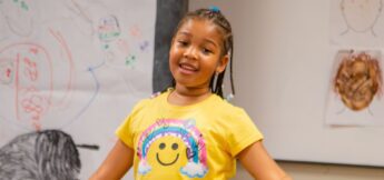 Young girl with braided hair wearing a yellow shirt with a smiley face stands in a classroom with drawings on the walls behind her.