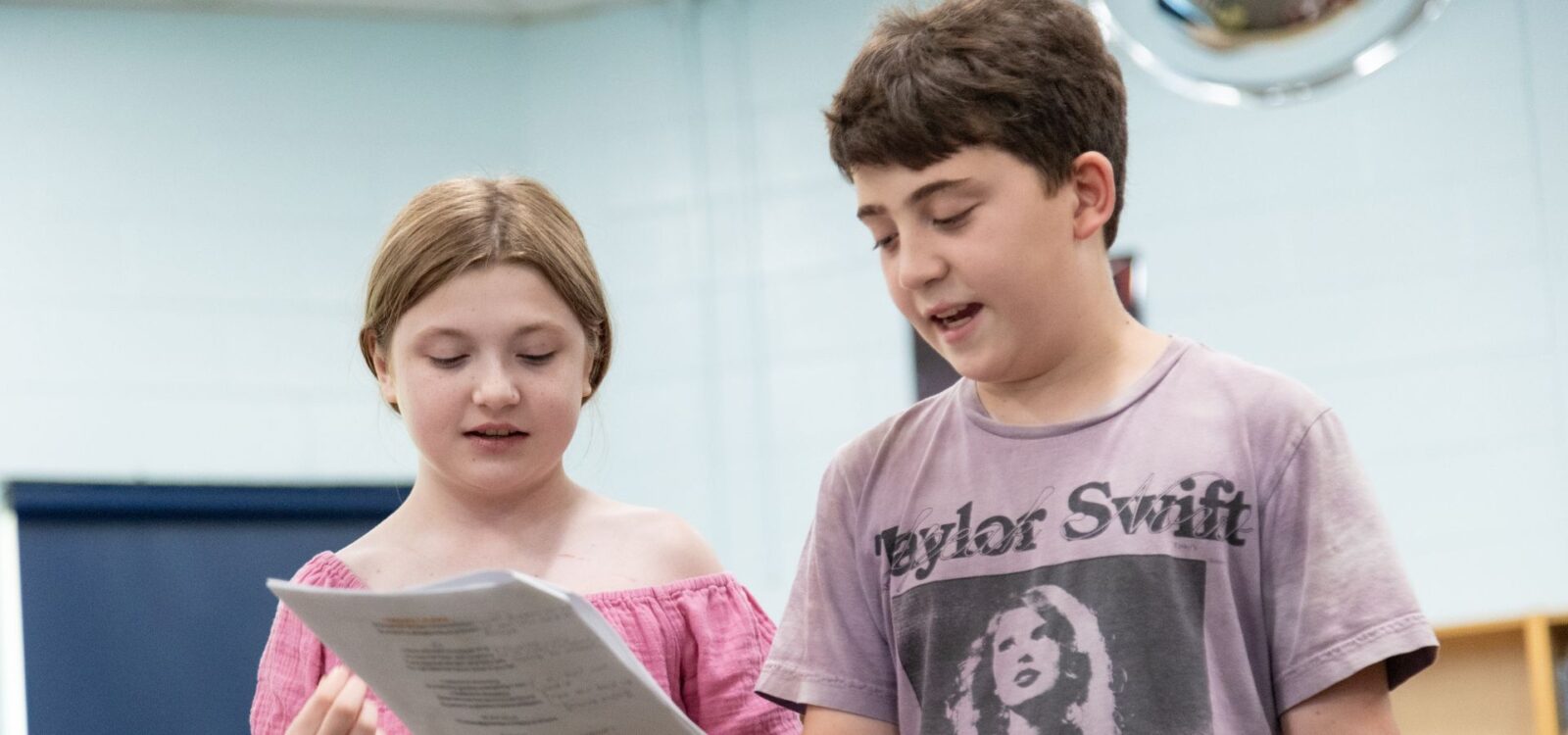 Two children stand indoors reading from a sheet of paper; one wears a pink shirt, the other wears a Taylor Swift T-shirt.