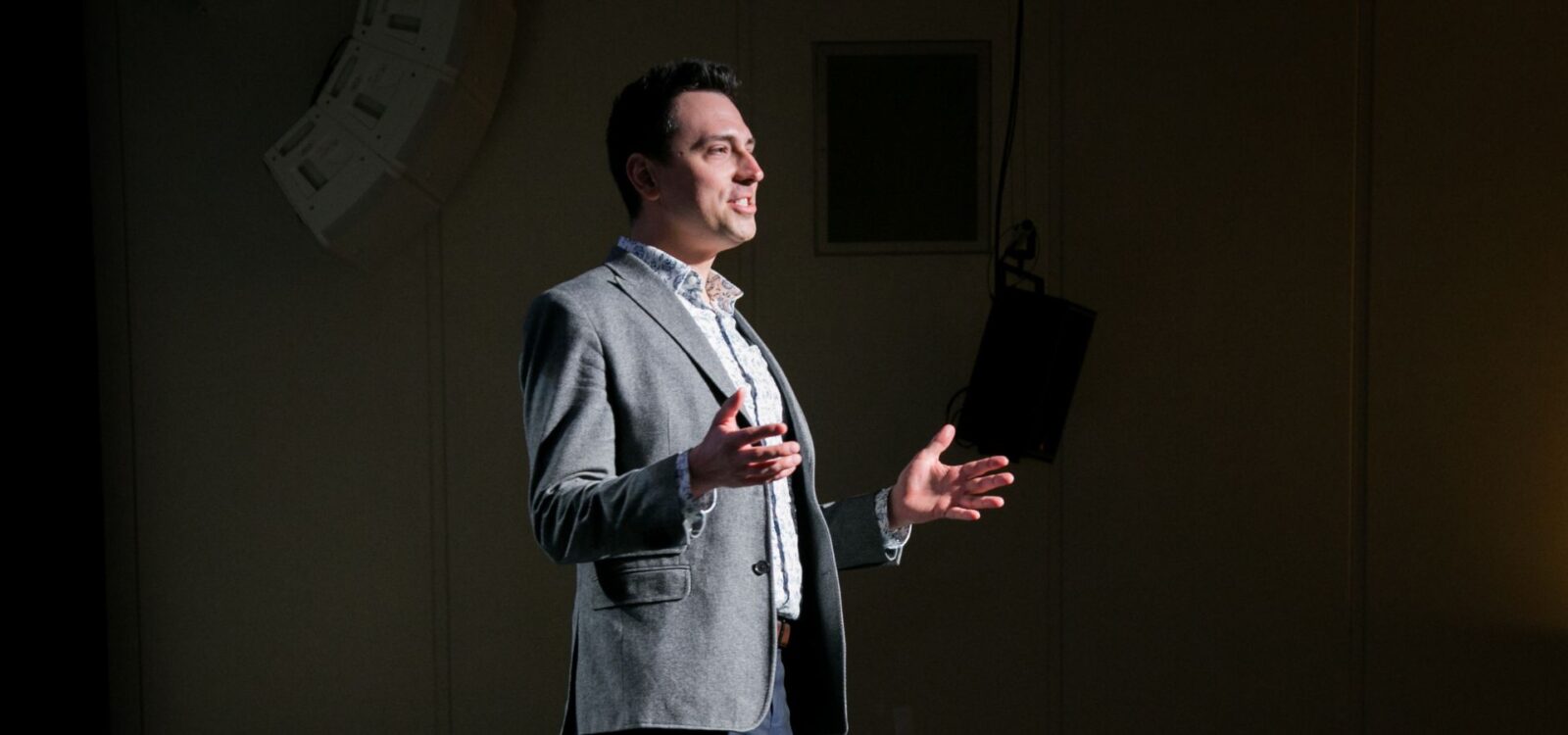A man in a gray blazer and white shirt stands on stage, speaking with open hands, illuminated by stage lighting.