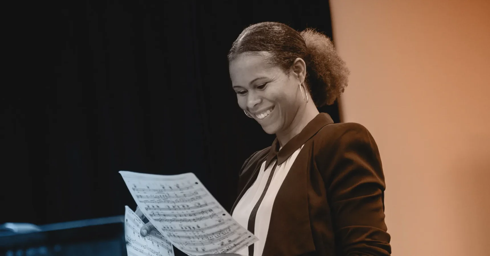 A woman smiling and looking at sheet music while standing indoors, dressed in a blazer and blouse, with a dark background behind her.