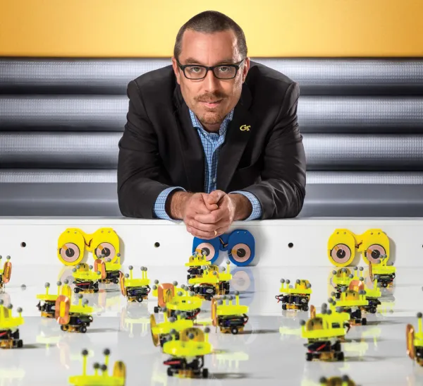 A man in a suit leans on a table covered with many small yellow robotic devices, with a yellow background and striped wall behind him.