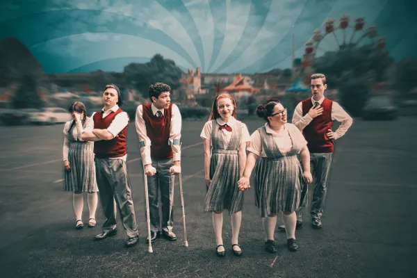 Six people in matching school uniforms stand in a parking lot, with a ferris wheel and carnival rides visible in the blurred background under a cloudy sky.