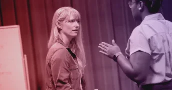 Two people are having a serious conversation indoors; one person is facing the camera, while the other is gesturing with their hands.