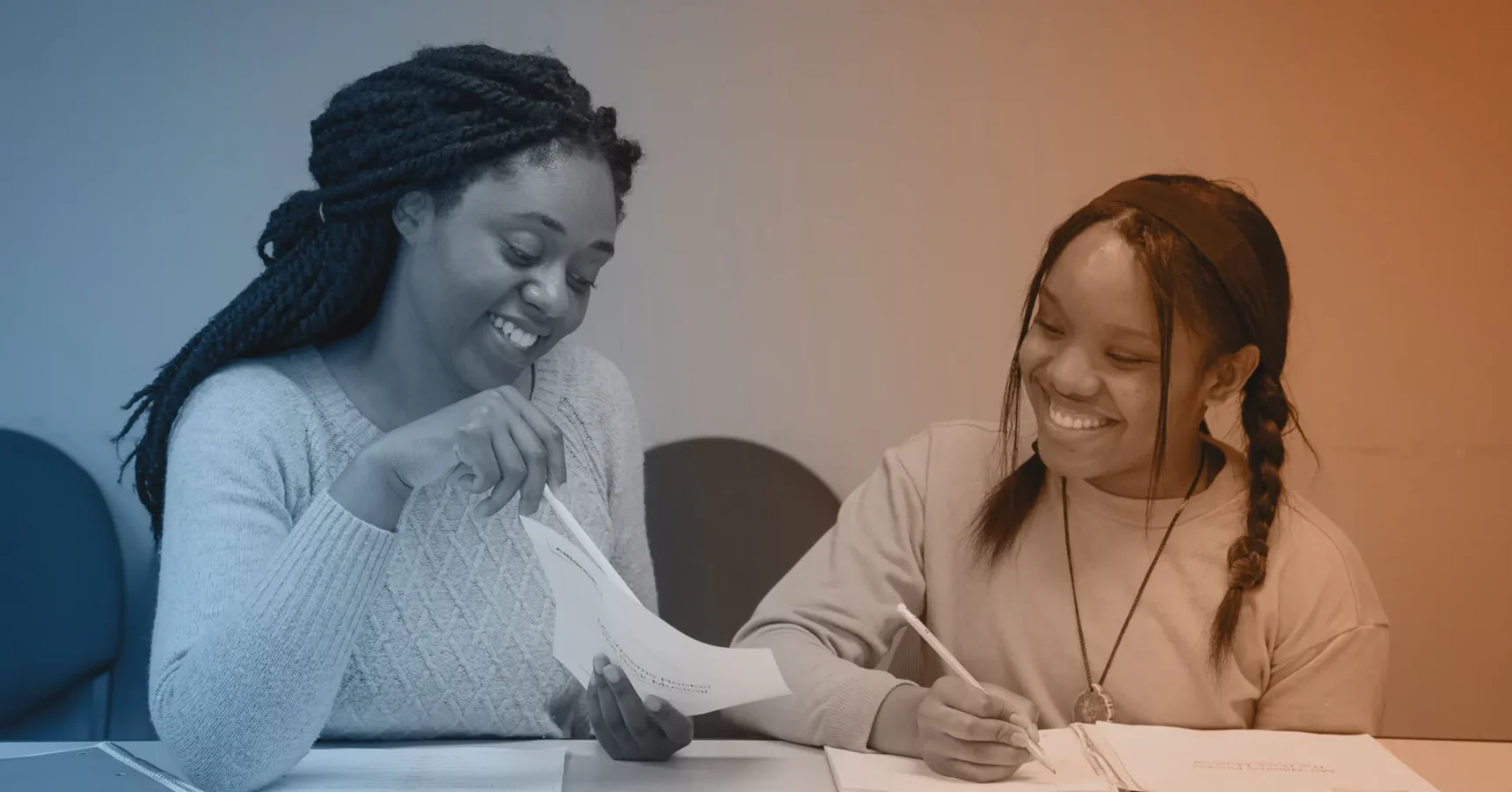Two women sit at a table smiling, one holding a piece of paper and the other writing with a pencil, appearing to engage in a collaborative or educational activity.