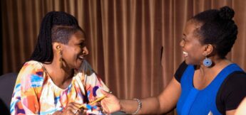 Two women sitting and smiling at each other, engaged in a lively conversation, with one holding a pencil. A brown curtain is in the background.