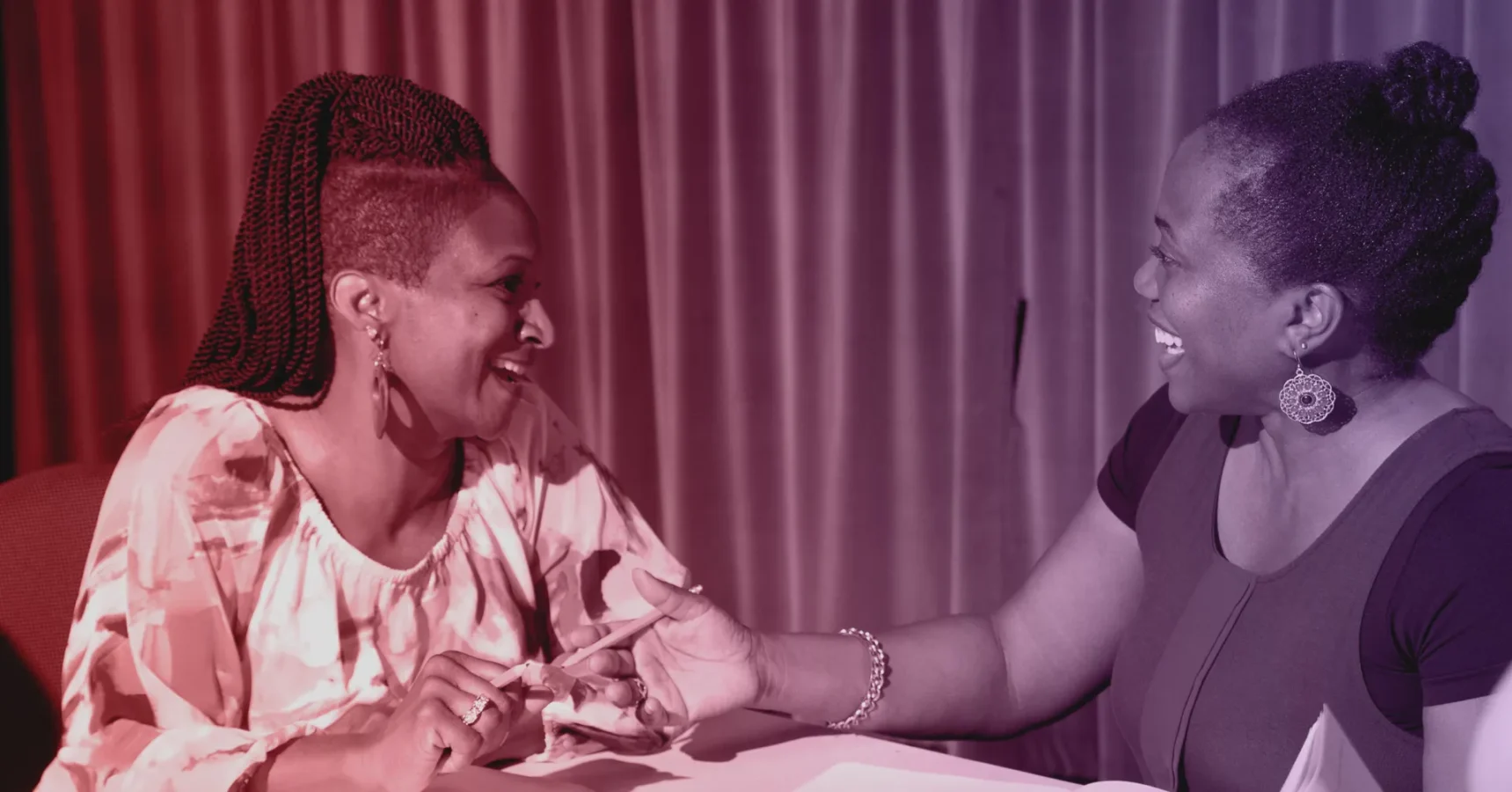 Two women sit at a table, facing each other and smiling while having a conversation, with curtains in the background and papers in front of them.