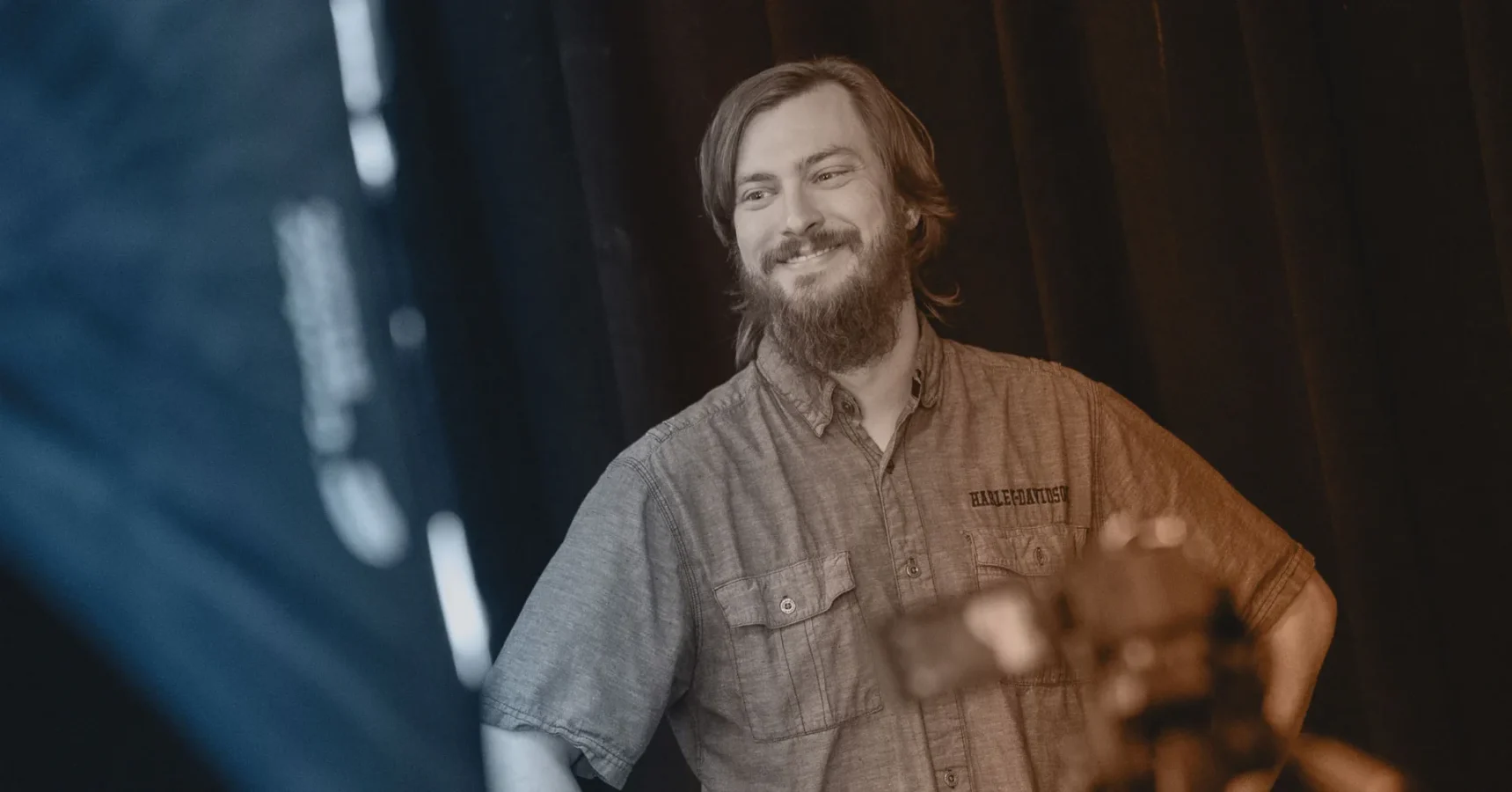 A bearded man wearing a button-up shirt stands in front of a dark background, smiling, with camera equipment visible in the foreground.