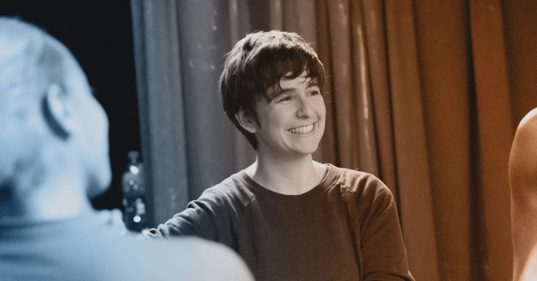 A person with short hair wearing a long-sleeve shirt is smiling, standing indoors near others with curtains in the background.