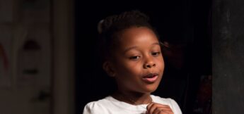 A young girl with dark hair in a bun, wearing a white shirt, stands indoors and looks slightly to the side, holding a necklace with one hand.