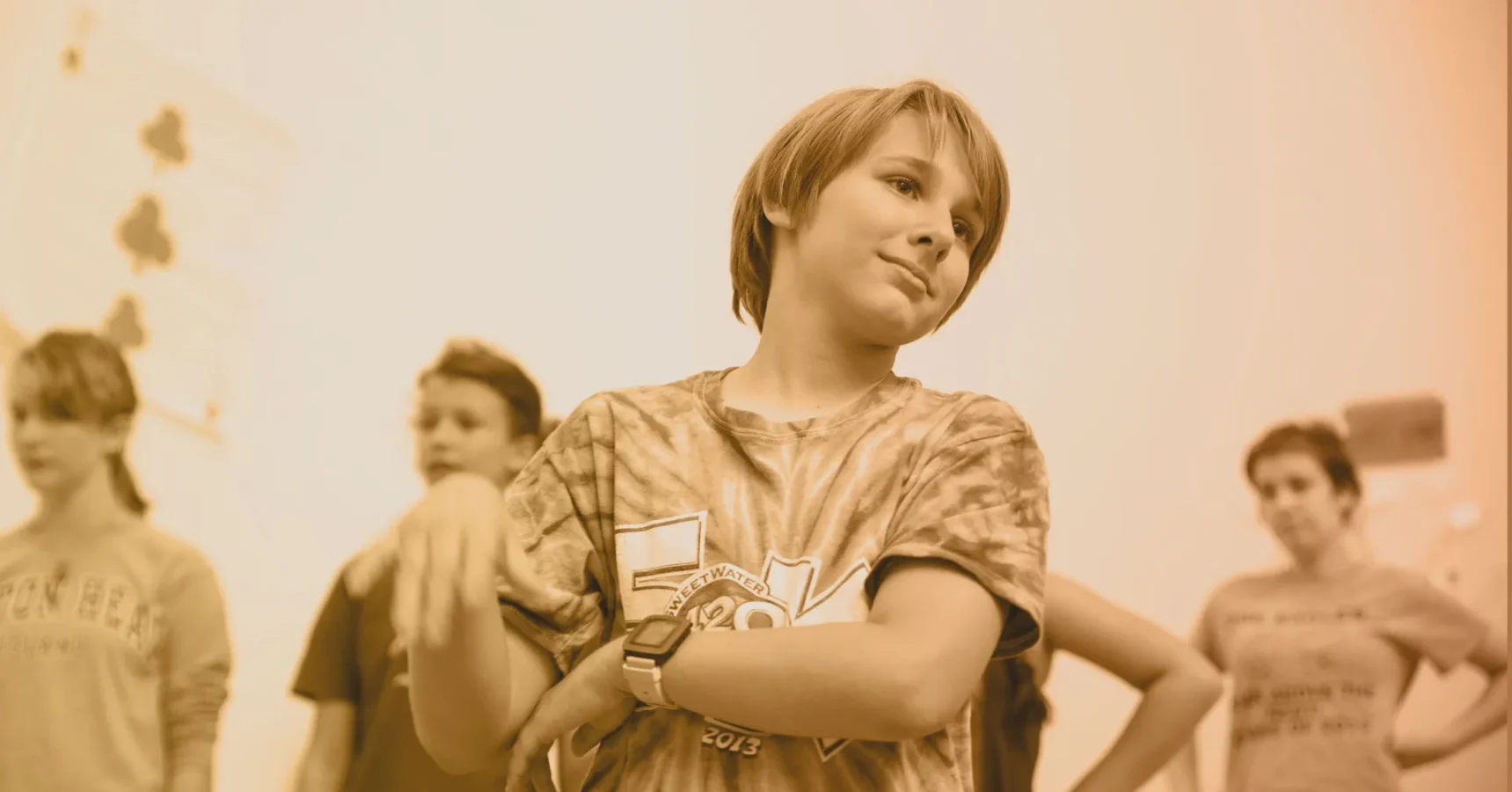 A group of children stand together indoors, with one child in the foreground crossing their arms and tilting their head, while others are visible in the background.