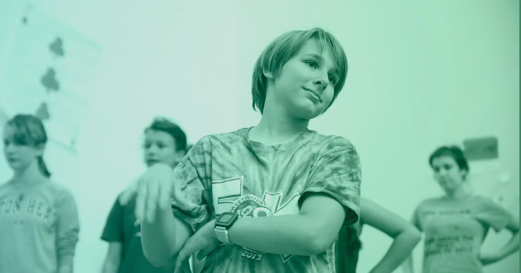A group of children stands indoors, with one child in the foreground crossing their arms and tilting their head, while others are blurred in the background.
