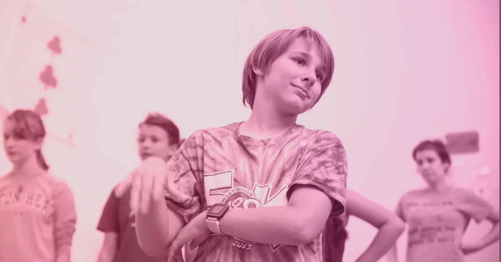 A group of children stand indoors, with one child in the foreground shrugging and making a nonchalant gesture, wearing a tie-dye shirt. The image has a pink tint.