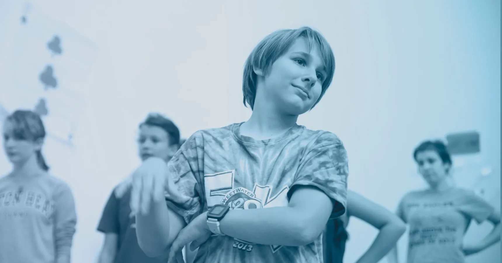 A group of young people stand together indoors; one person in the foreground has arms crossed and a neutral expression.