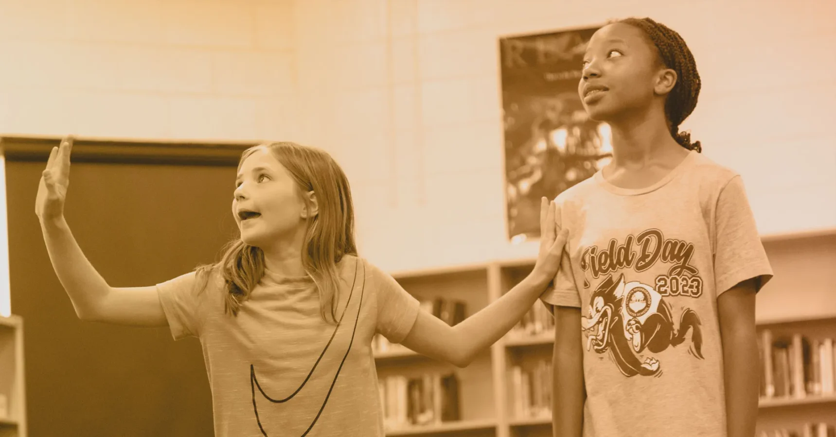 Two children stand in a room with bookshelves; one gestures animatedly with both hands while the other looks upward.