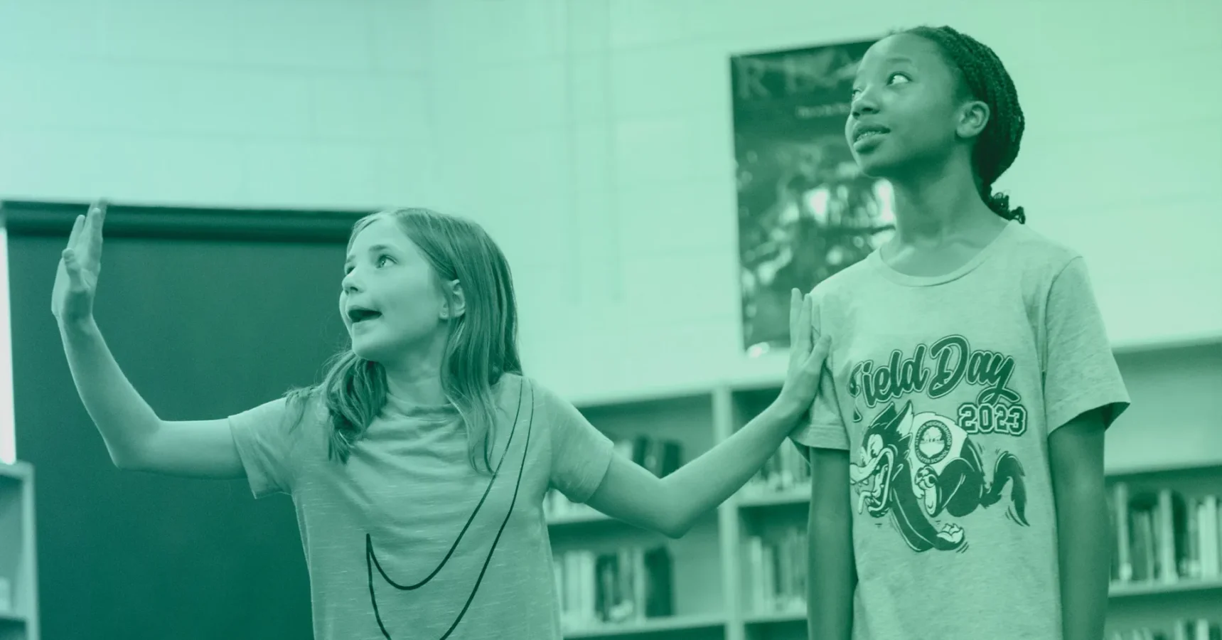 Two girls stand in a classroom; one gestures with her arms raised while the other looks upward. Shelves and a poster are visible in the background.