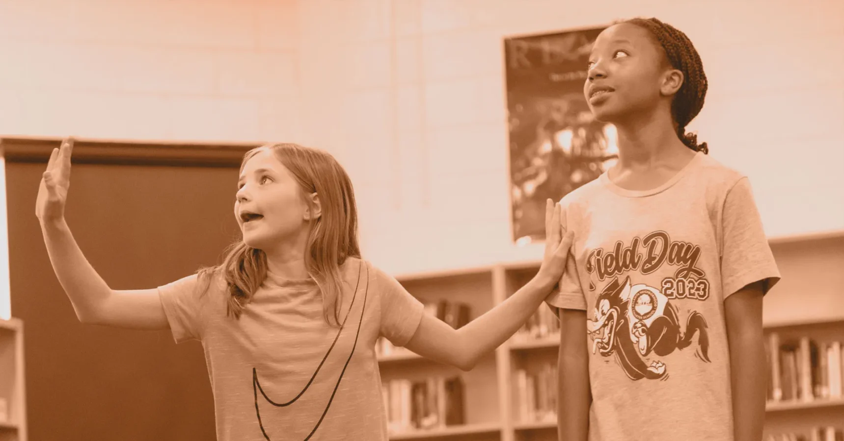 Two girls stand in a library; one gestures energetically while the other looks up with a neutral expression.