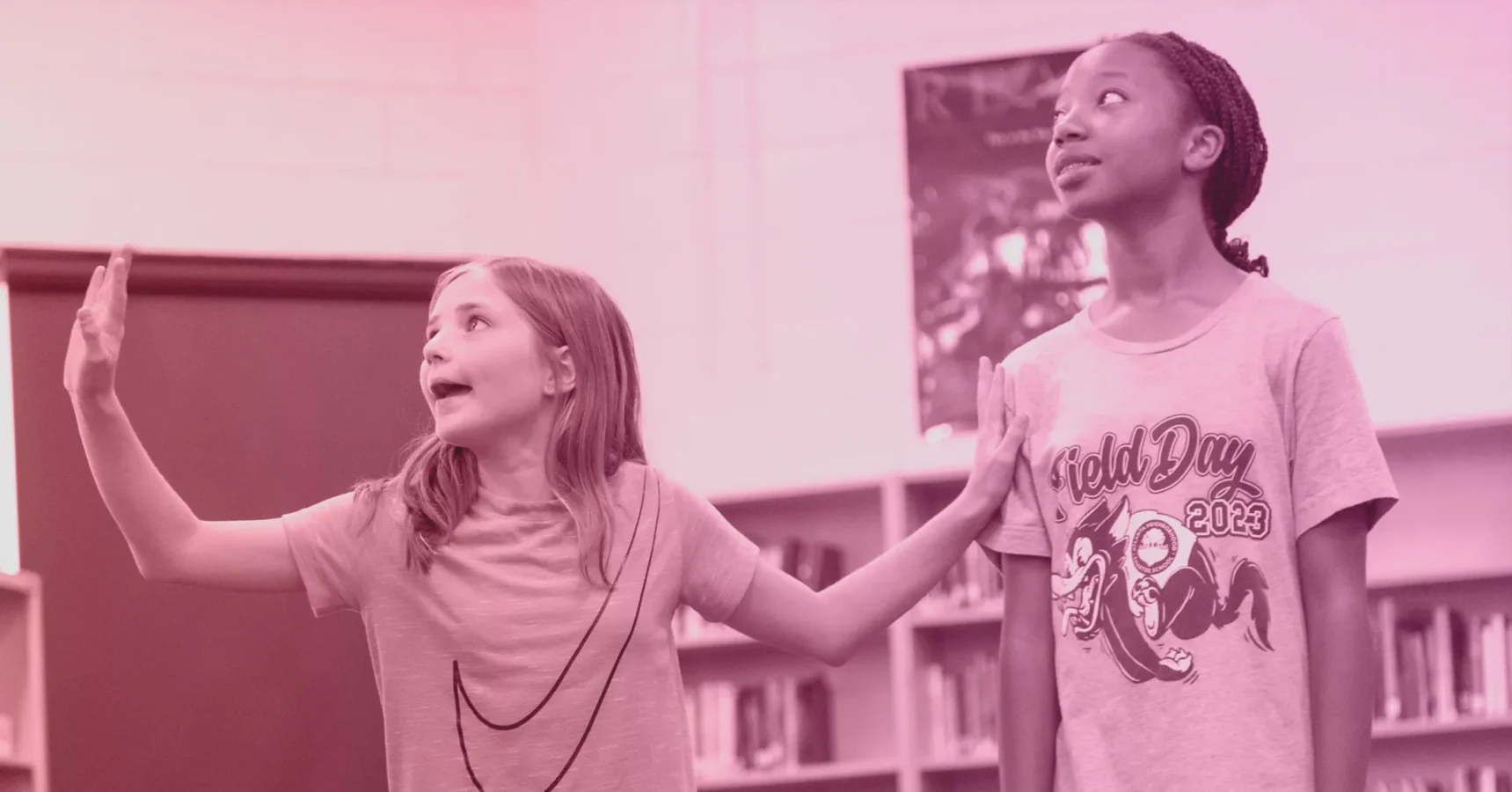 Two girls stand in a library; one raises her hands while speaking, the other looks up. Bookshelves and a poster are visible in the background.