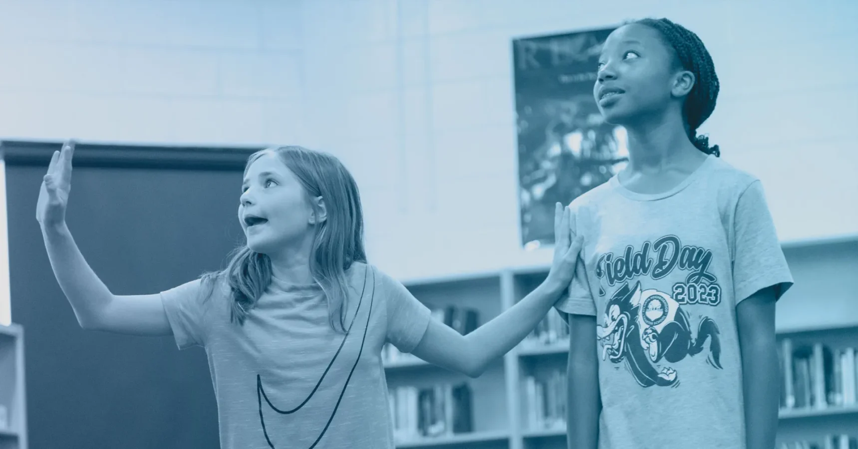 Two girls stand in a library; one gestures animatedly with her hand raised while the other looks upward with a neutral expression.