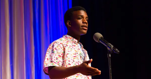 A person in a colorful shirt speaks into a microphone on stage, gesturing with one hand. Blue and white curtains are in the background.