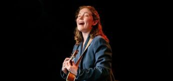 A person with long hair plays a ukulele and sings on stage against a dark background.