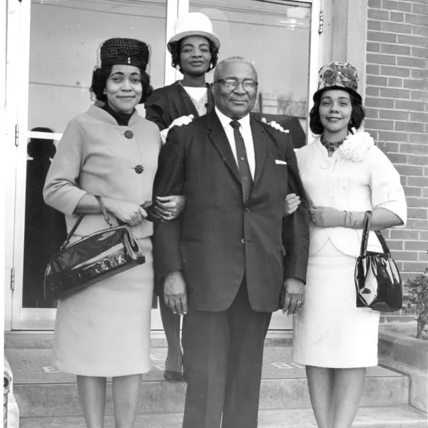 A man in a suit stands arm-in-arm with three women in formal attire and hats, posing on the steps outside a building.