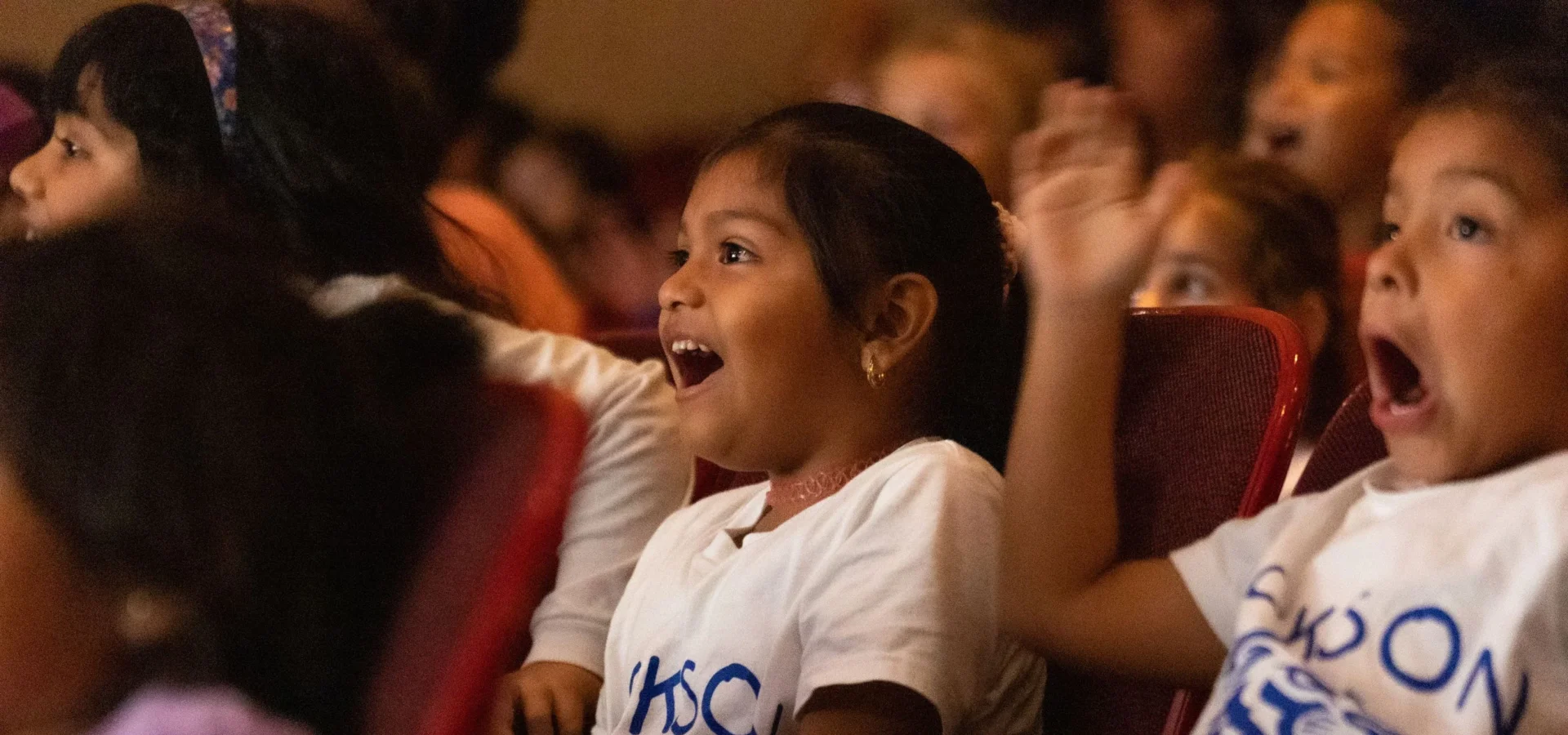 Young children sit in theater seats, watching a performance with excited and surprised expressions on their faces.