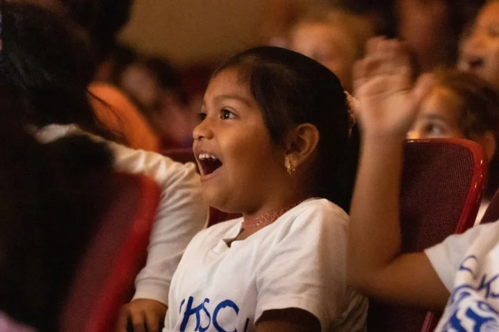 Young children sit in theater seats, watching a performance with excited and surprised expressions on their faces.