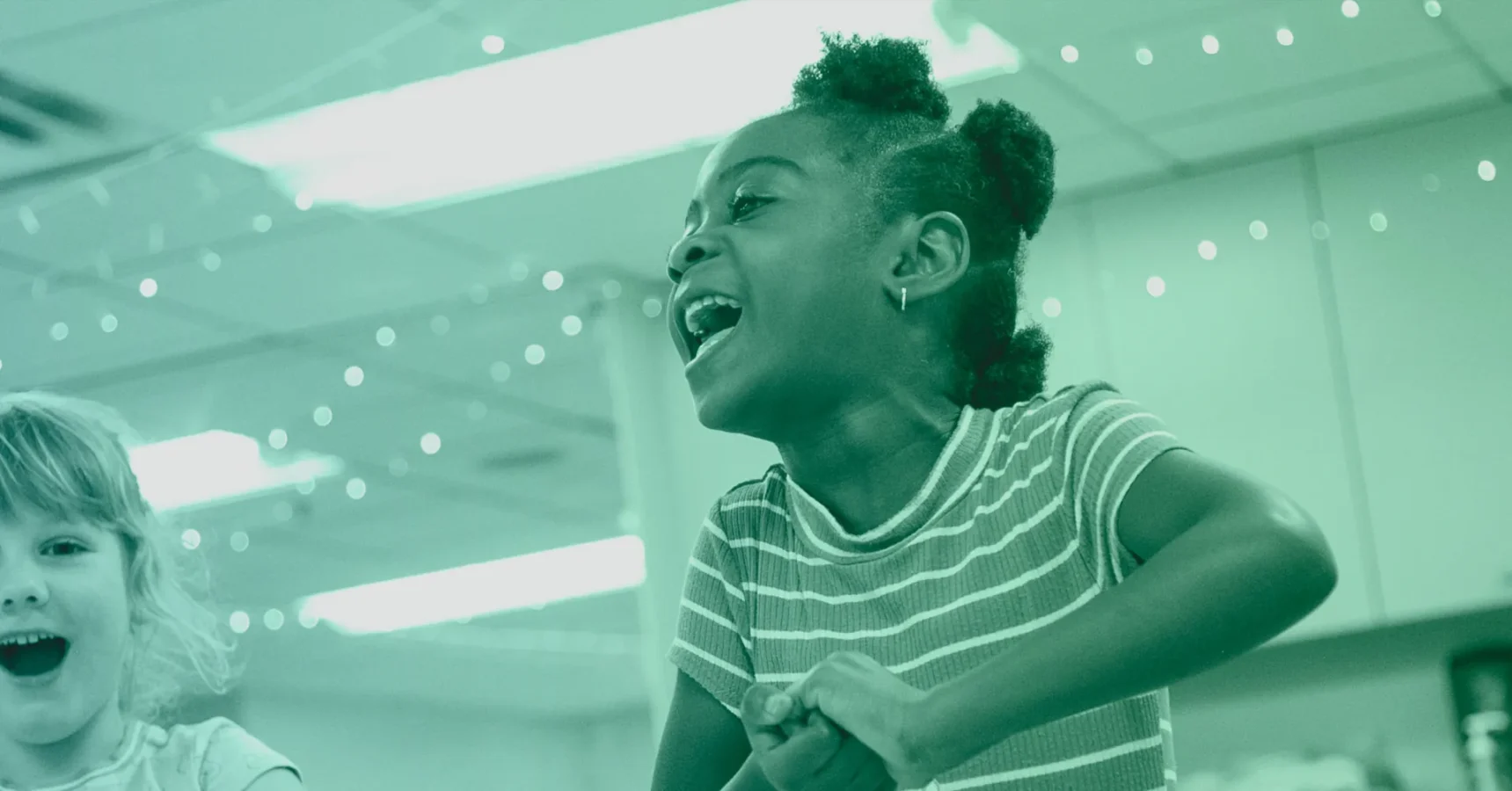 Two children indoors, one girl in a striped shirt smiles and laughs, appearing to be playing or interacting energetically, with lights and ceiling panels in the background.