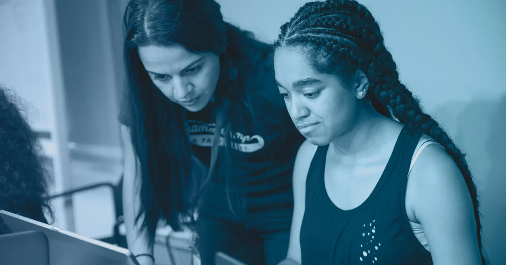 Two young women look at a laptop screen together; one is seated and typing while the other leans over to observe.