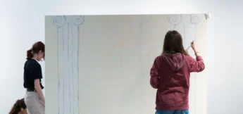 Two people stand in front of a large canvas, drawing outlines of classical columns with faint lines visible on the canvas at high school technical theater camp.