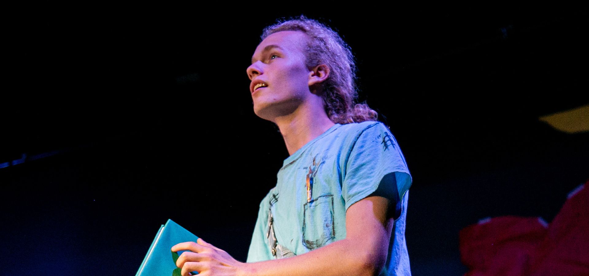 A person with curly hair wearing a light blue shirt holds a turquoise folder while looking upward on a dimly lit stage.