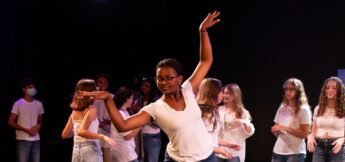 A group of young people in white shirts and jeans perform on stage, with one person dancing in the foreground and others clapping or dancing in the background.