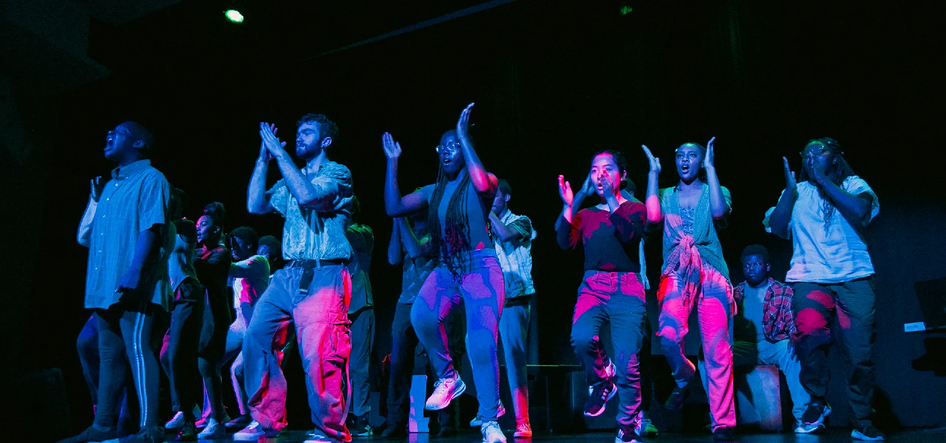 A group of people performs on stage, clapping and dancing under blue and pink stage lighting.