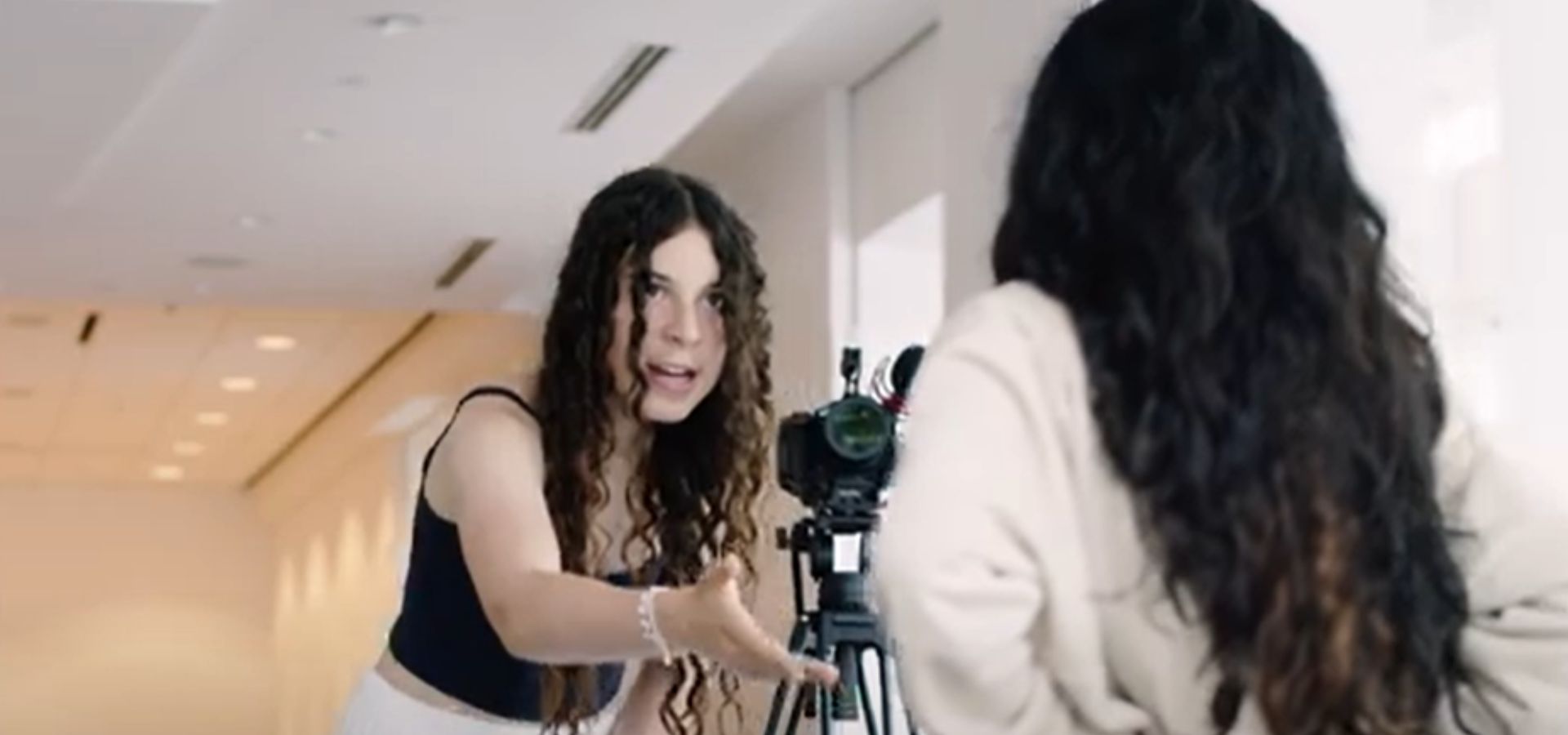 A woman with long curly hair gestures while speaking to another woman in front of a camera on a tripod in a hallway.