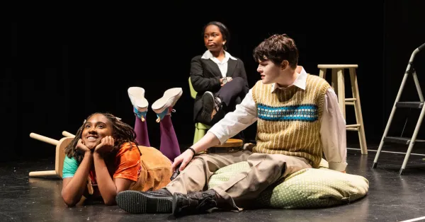 Three people are on a stage; one lies on the floor smiling, another sits cross-legged with an arm outstretched, and a third sits on a stool in the background.