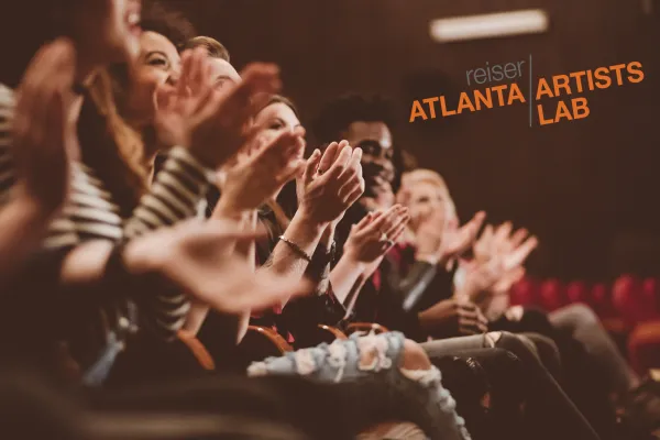 A group of people sit in a theater audience, clapping, with the words "Atlanta Artists Lab" displayed on the wall in the background.