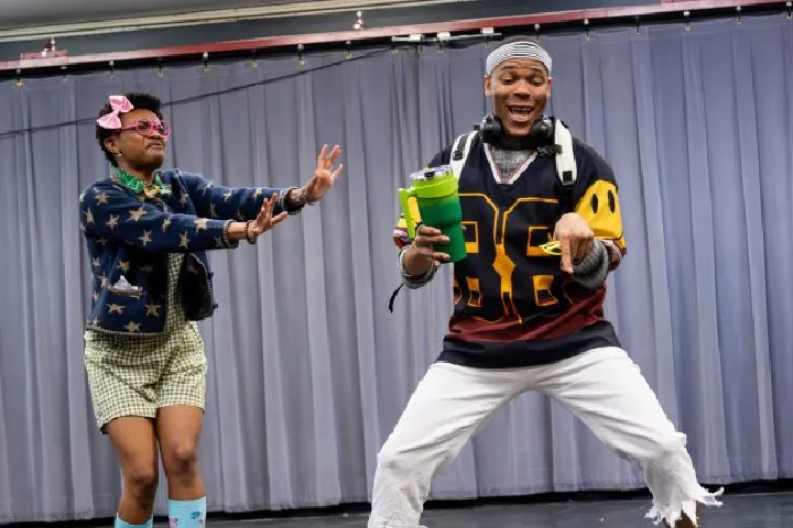 Two people in colorful, quirky outfits perform on stage in front of a gray curtain; one gestures with hands while the other holds a green water bottle and dances.
