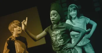 Three children perform on stage, with one child in the foreground raising a finger and the other two in the background, all appearing to be mid-dance or acting.