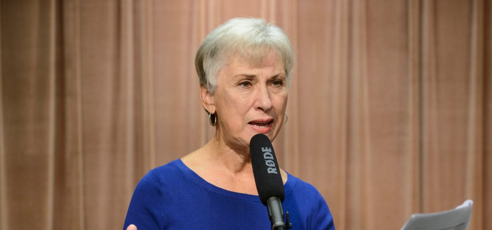 An older woman with short gray hair speaks into a Røde microphone while holding papers, standing in front of a brown patterned background.