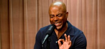 A person passionately sings or speaks into a microphone, eyes closed, wearing a navy blue shirt, with a tan curtain background.