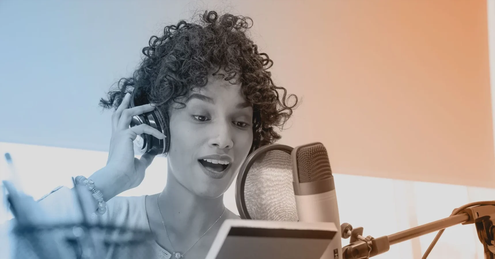 A person wearing headphones is speaking or singing into a professional microphone while reading from a tablet or book in a recording studio.