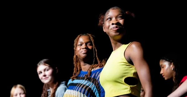 Five young women stand on stage, some smiling, under a spotlight with a black background.