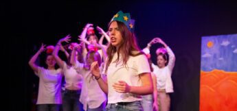 A young woman wearing a blue crown with yellow stars performs on stage, with several people in white shirts and red headbands posing in the background.