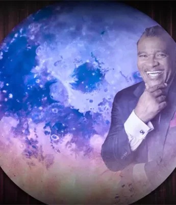 A man in a suit smiles with his hand on his chin, standing in front of an artistic, colorful moon backdrop.