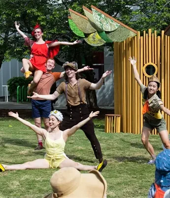 A group of performers in colorful costumes pose with arms outstretched on a grassy outdoor stage in front of an audience.