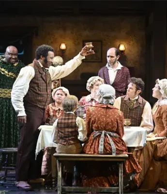 A group of actors in period costumes sit and stand around a dining table on stage, with one man raising a glass in a toast.