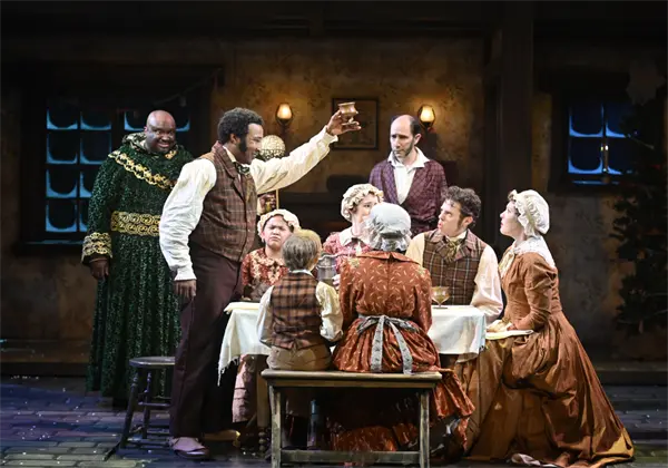 A group of actors in period costumes sit and stand around a dining table on stage, with one man raising a glass in a toast.