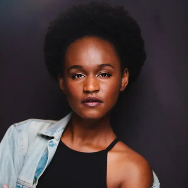A woman with a natural afro hairstyle wears a denim jacket over a black top, standing against a dark background and looking directly at the camera.