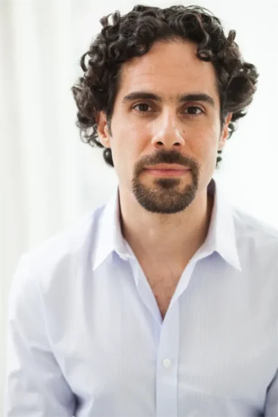 A man with curly dark hair, a goatee, and mustache, wearing a light-colored button-up shirt, looks directly at the camera against a bright, neutral background.